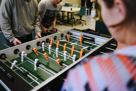 Foto von Studierenden, die am Kicker in der BibLounge am Standort Sonnenstraße spielen. Im Hintergrund stehen Tische und Stühle__Photo of students playing table football in the BibLounge at campus Sonnstraße, with tables and chairs in the background.