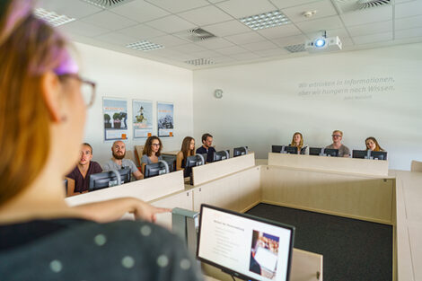 Photo of eight students sitting at PCs in a seminar room. They are listening to a young woman in the front left corner of the picture.__Eight students are sitting at PCs in a seminar room listening to a young woman in the front left corner of the picture.