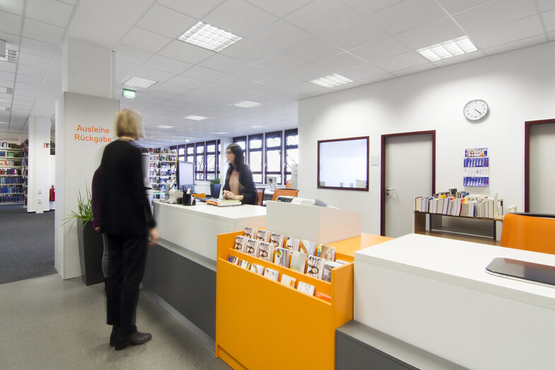 Foto zweier Studierender im Gespräch mit einer Mitarbeiterin der Bibliothek an der Ausleihtheke__Photo of two students in conversation with a female member of library staff at the service counter