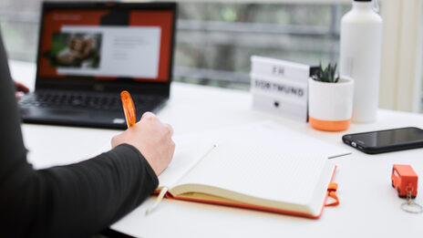 Photo of a workplace with a laptop. In the foreground you can see the arm of a person writing something in a notebook.