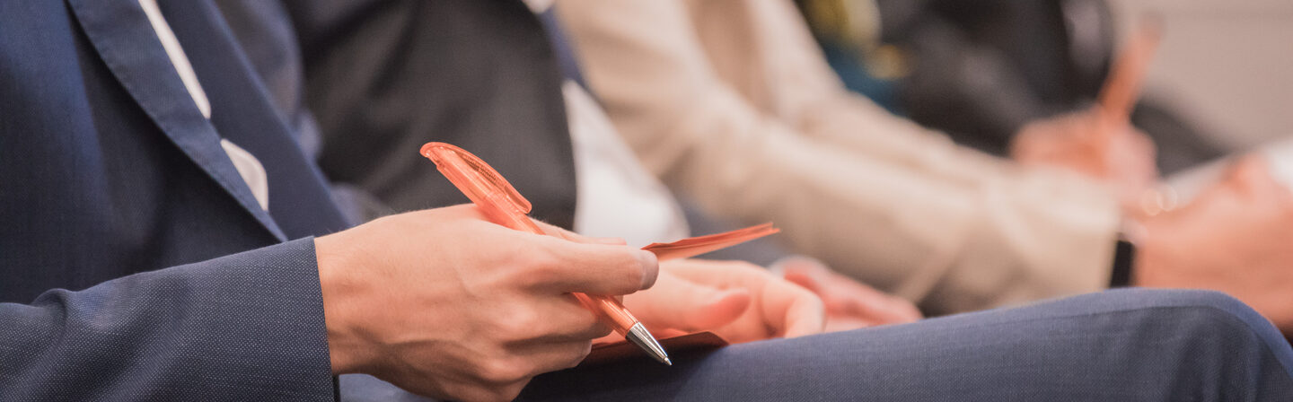 Photo of a row of people sitting at an event. A man holding pen and material in hand. __ Row of people sitting at an event, man holding pen and material in hand.