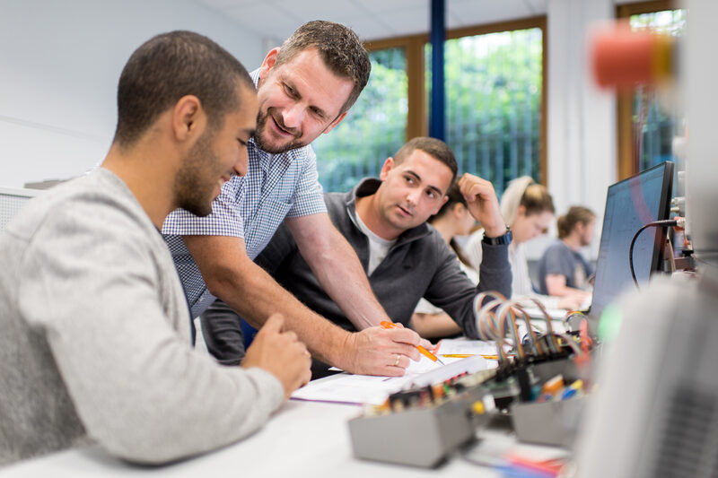 Photo from the electronics and automation laboratory. A professor helps two students at workstations with electronic experimental setups. Other students in the background.