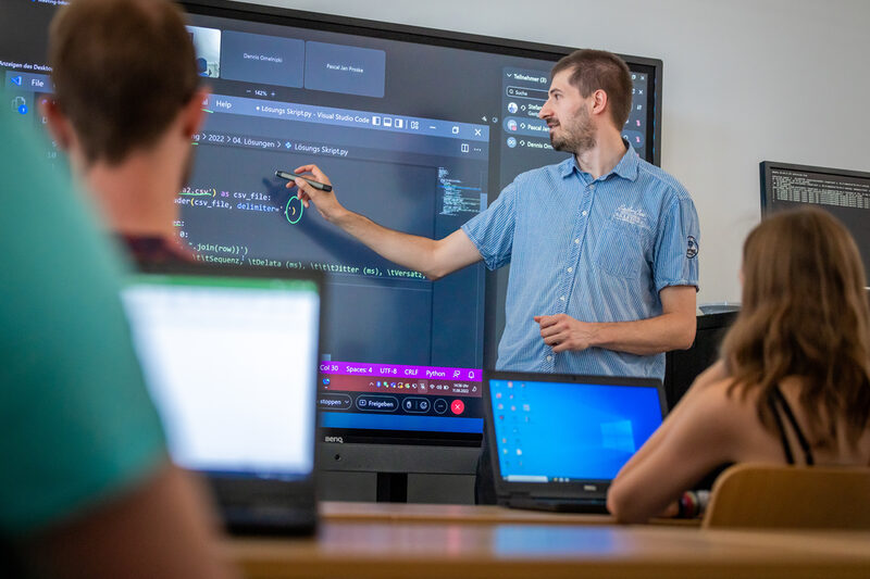 Foto von einer Vorlesungssituation in einem Seminarraum. Vorne an einem digitalen Board steht ein Mann und erklärt etwas in einem Code.