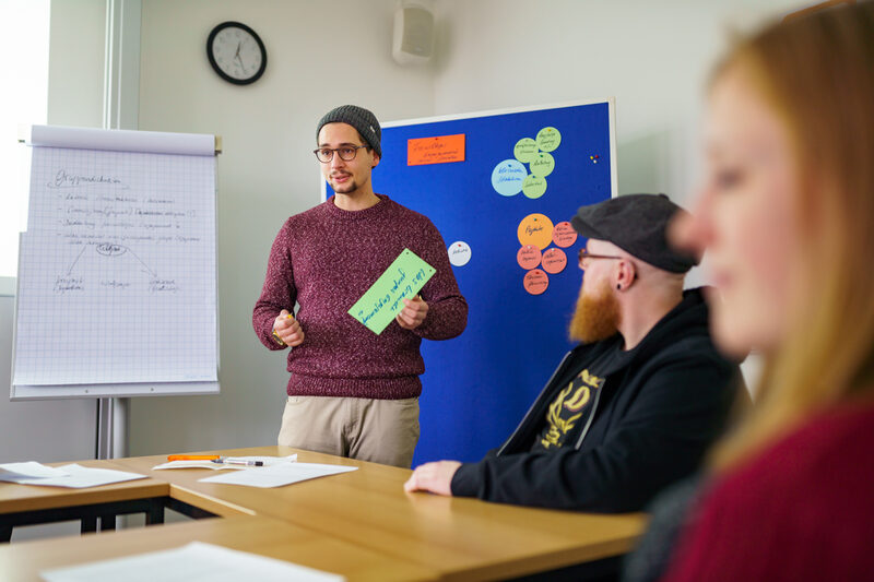 Photo of a member of the ethnography working group. The young man is standing in the room holding a green piece of paper. Behind him is a flipchart and a pinboard with colorful notes pinned to it. Two people are sitting on the right.