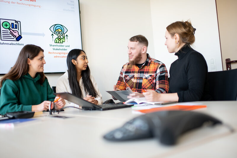 Foto von Studierenden, die an einem Tisch sitzen und sich unterhalten. Vor ihnen steht ein Laptop und links hinter ihnen ist eine Beamerleinwand.