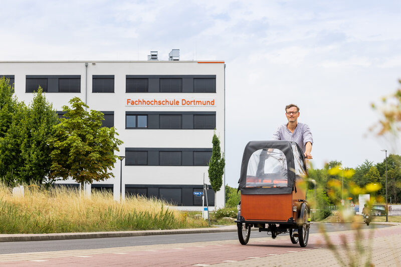 Foto von einem Mann auf einem Lastenfahrrad, der auf dem Radweg von einem Gebäude wegfährt. Auf dem Gebäude ist der Schriftzug "Fachhochschule Dortmund" zu sehen.