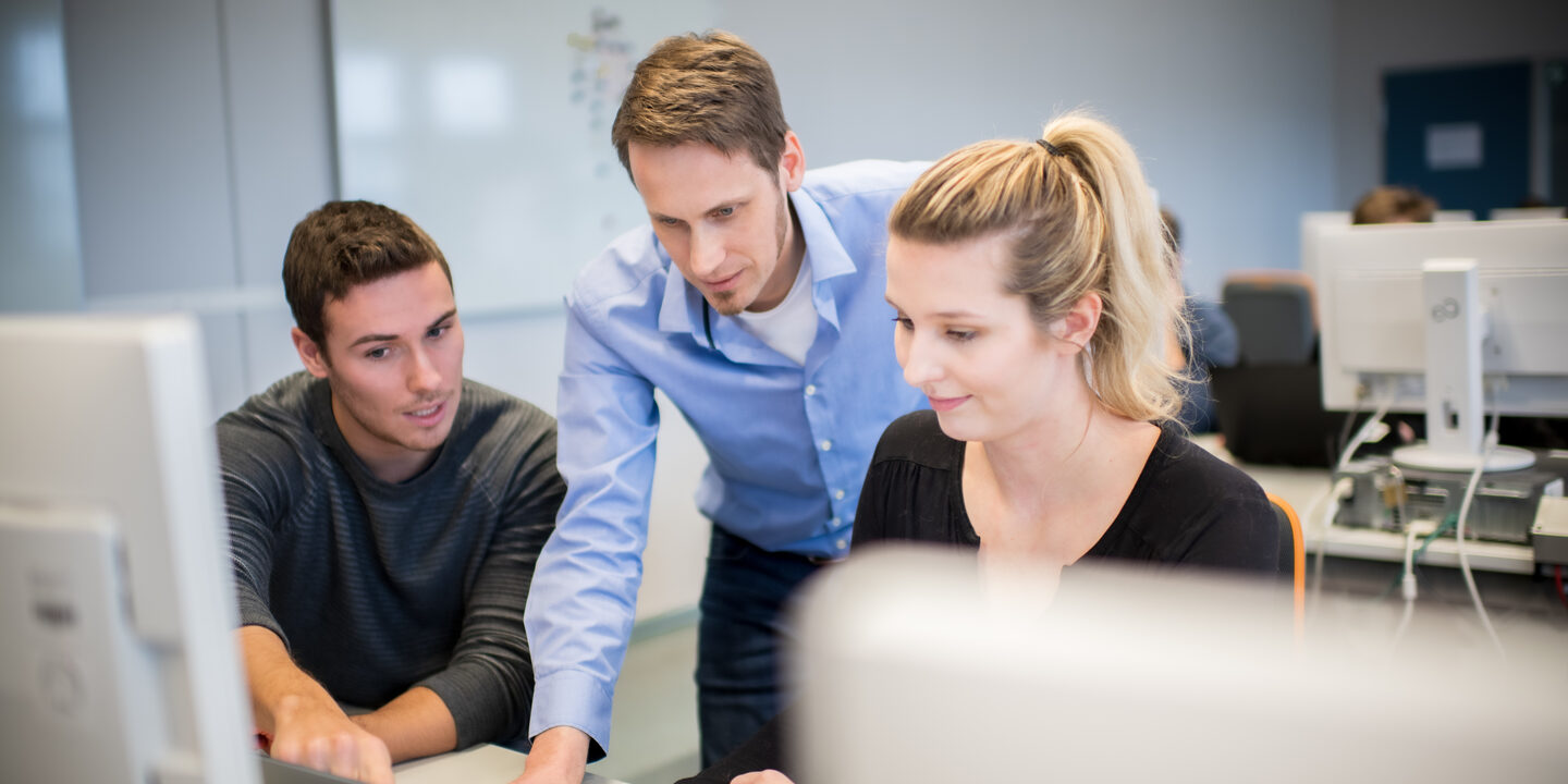 Foto eines Lehrenden im Computerraum, der zwischen einem Studenten und einer Studentin steht und beiden etwas am Laptop erklärt.  __ <br>The professor stands between a male student and a female student in the computer room and explains something to both of them on the laptop.