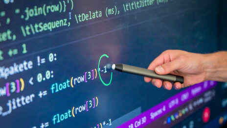 Photo of a section of a digital board on which a code can be seen. Someone is holding a pen pointing to a spot.