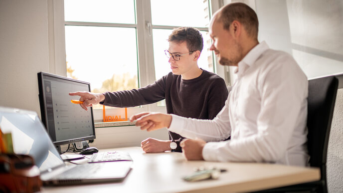 Foto von 2 Beschäftigten, die an einem Arbeitsplatz sitzen und etwas am Rechner besprechen.