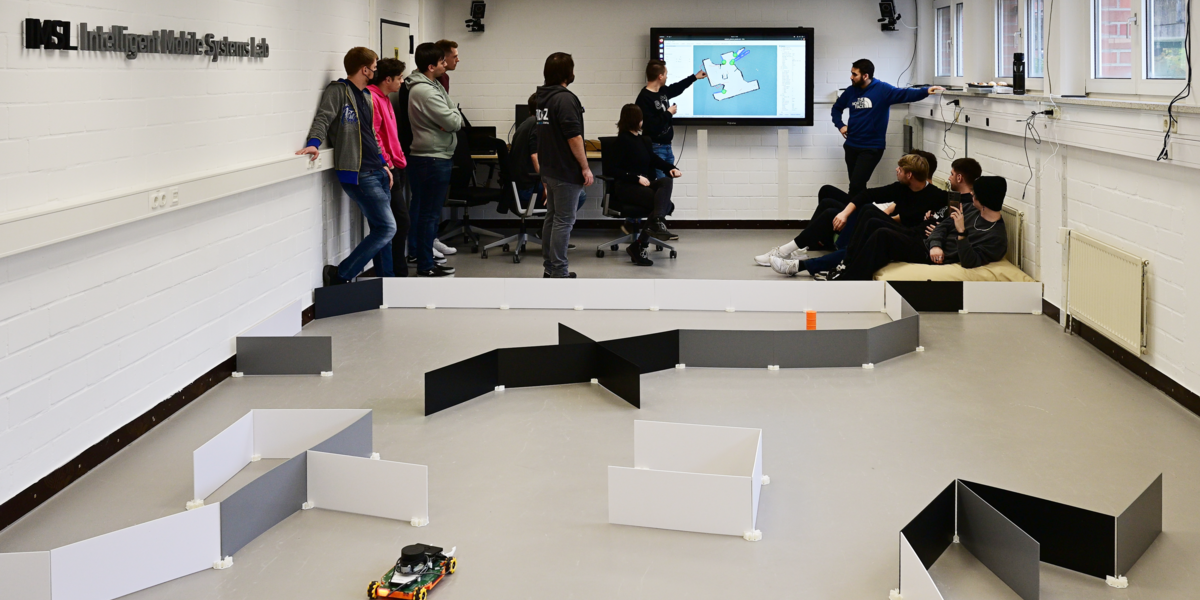 In the background, a group of 12 students and the lecturer are standing and sitting in front of a television on a wall. A map created by a robot can be seen on the television. A student explains details on the map while pointing to the TV with his left hand. In the foreground there is a course made of upright plastic plates in which an EduRob with an open gripper and a model of a hazardous materials container can be seen.