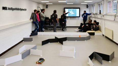 In the background, a group of 12 students and the lecturer are standing and sitting in front of a television on a wall. A map created by a robot can be seen on the television. A student explains details on the map while pointing to the TV with his left hand. In the foreground there is a course made of upright plastic plates in which an EduRob with an open gripper and a model of a hazardous materials container can be seen.