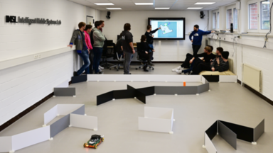 Im Hintergrund ist eine Gruppe von 12 Studierenden und der Dozent stehen und sitzen vor einem Fernseher an einer Wand. Auf dem Fernseher ist eine von einem Roboter erstellte Karte zu sehen. Ein Student erklärt Details auf der Karte und zeigt dabei mit der linken Hand auf den Fernseher. Im Vordergrund ist Parcours aus stehenden Kunststoffplatten zu sehen in dem ein EduRob mit offenem Greifer und ein Modell eines Gefahrenstoffbehälters steht.