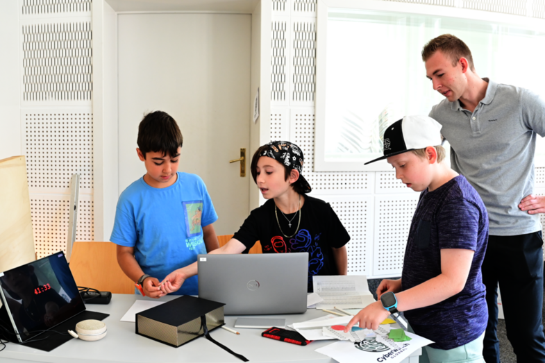 Three students stand around a table with a notebook and various utensils to solve the "digital rescue" puzzle. A tablet displays the remaining time of 41:33 and an employee of the UAS gives a hint to solve the puzzle.