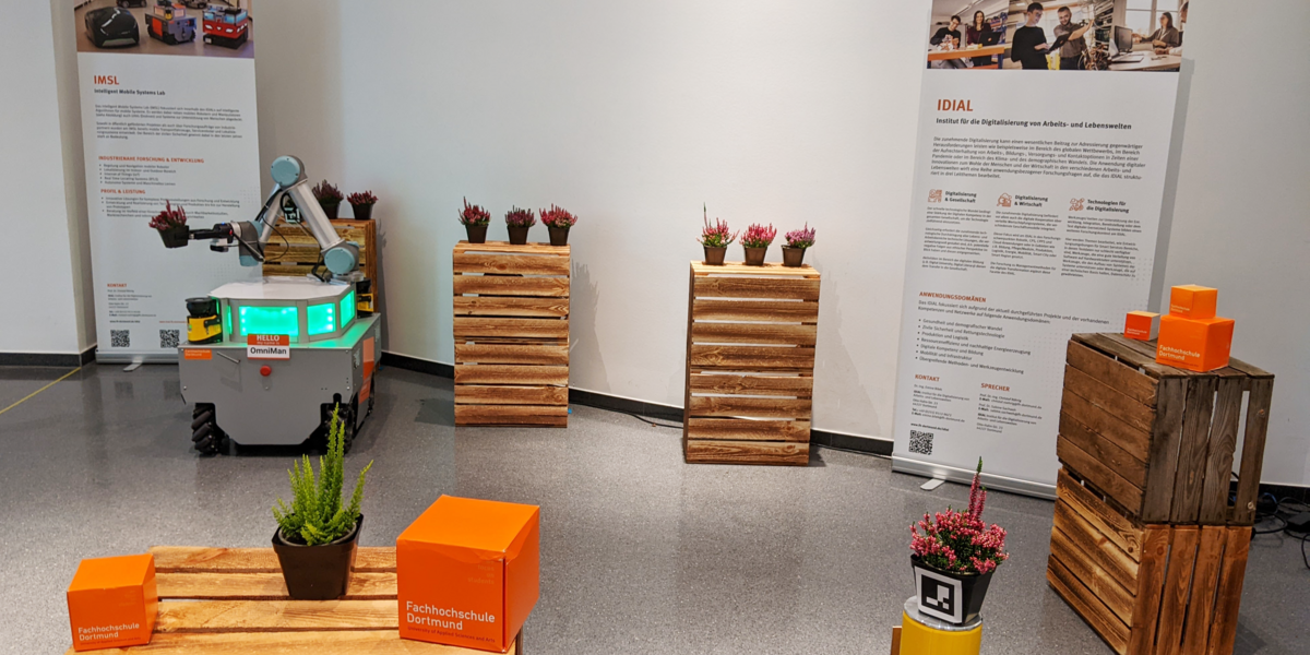 The mobile manipulator OmniMan stands in the Dortmunder U between towers of wine crates on which plant pots with heather are placed. OmniMan transports a plant pot by holding it in its two-finger gripper. In the background are roll-ups from IDiAL and the Intelligent Mobile System Lab. In the foreground, UAS cubes and a heather plant are set up on a crate tower.