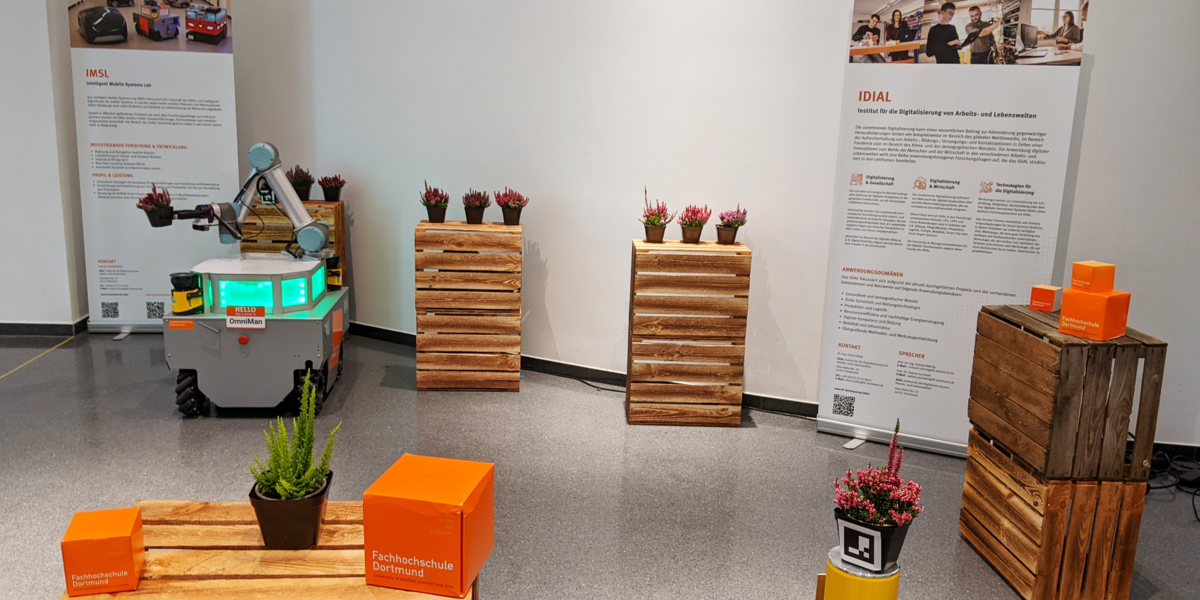 Der mobile Manipulator OmniMan steht im Dortmunder U zwischen Türmen aus Weinkisten, auf denen Pflanztöpfe  mit Heidekraut stehen. OmniMan transportiert einen Pflanztopf indem er ihn in seinem Zwei-Finger-Greifer hält. Im Hintergrund stehen Rollups vom IDiAL und des Intelligent Mobile System Labs. Im Vordergrund sind FH-Würfel und ein Heidekrauf auf einem Kistenturm aufgestellt.