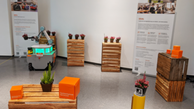 Der mobile Manipulator OmniMan steht im Dortmunder U zwischen Türmen aus Weinkisten, auf denen Pflanztöpfe  mit Heidekraut stehen. OmniMan transportiert einen Pflanztopf indem er ihn in seinem Zwei-Finger-Greifer hält. Im Hintergrund stehen Rollups vom IDiAL und des Intelligent Mobile System Labs. Im Vordergrund sind FH-Würfel und ein Heidekrauf auf einem Kistenturm aufgestellt.