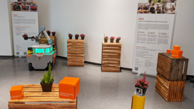 The mobile manipulator OmniMan stands in the Dortmunder U between towers of wine crates on which plant pots with heather are placed. OmniMan transports a plant pot by holding it in its two-finger gripper. In the background are roll-ups from IDiAL and the Intelligent Mobile System Lab. In the foreground, UAS cubes and a heather plant are set up on a crate tower.