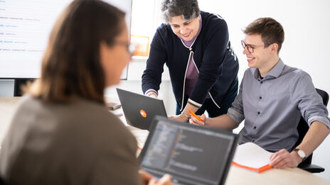 Photo of a woman and a colleague discussing something on a laptop, the woman explains something. Sitting opposite, another woman is blurredly recognizable at her laptop.