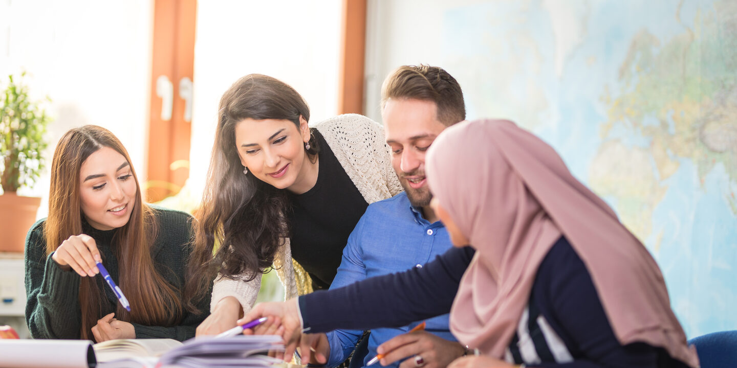 Eine Gruppe aus vier Studierenden aus verschiedenen Herkunftsländern beugt sich gemeinsam am Schreibtisch über Bücher.