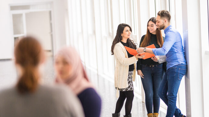 Foto von fünf internationalen Studierenden, von denen sich zwei im Vordergrund unterhalten und die anderen drei gemeinsam in eine orangefarbene Mappe sehen.
