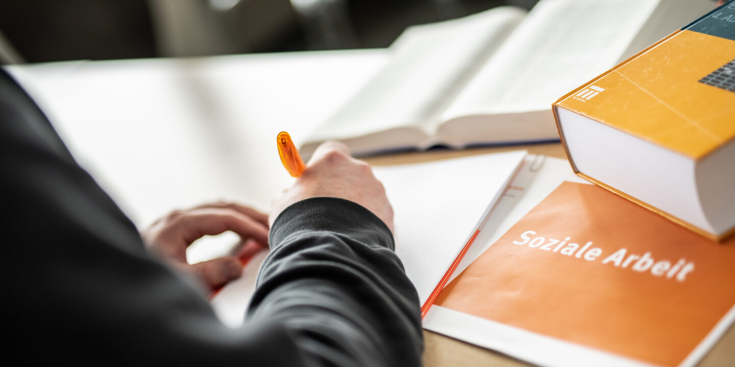 Foto einer Person im Anschnitt, die etwas mit einem orangefarbenen Stift auf einen Block schreibt. Daneben liegen Bücher und ein Heft mit Aufschrift "soziale Arbeit".