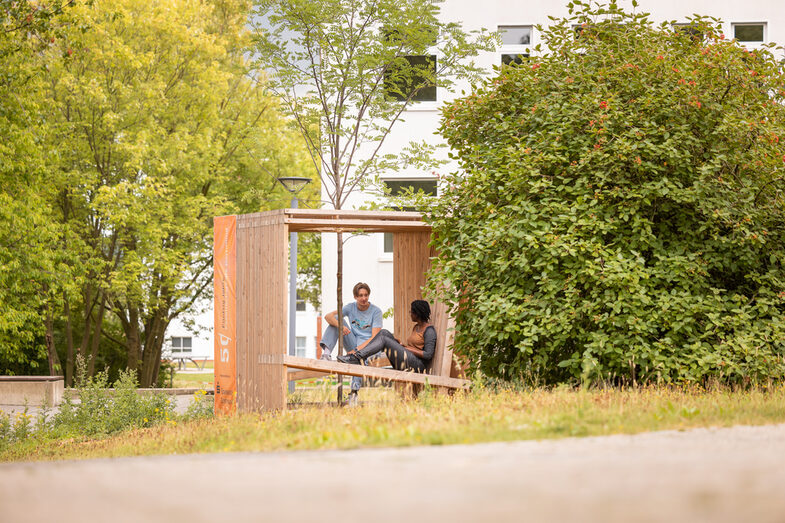 Photo of 2 students sitting and talking in a wooden cube equipped with seating. A tree is planted in the center of the cube.