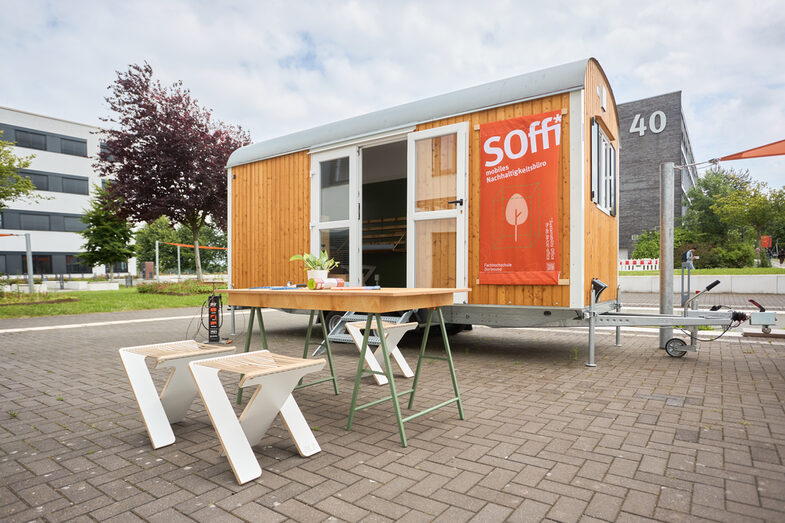You can see a construction trailer with a poster saying "Soffi mobile sustainability office"
In front of the trailer you can see a table with a few things on it.