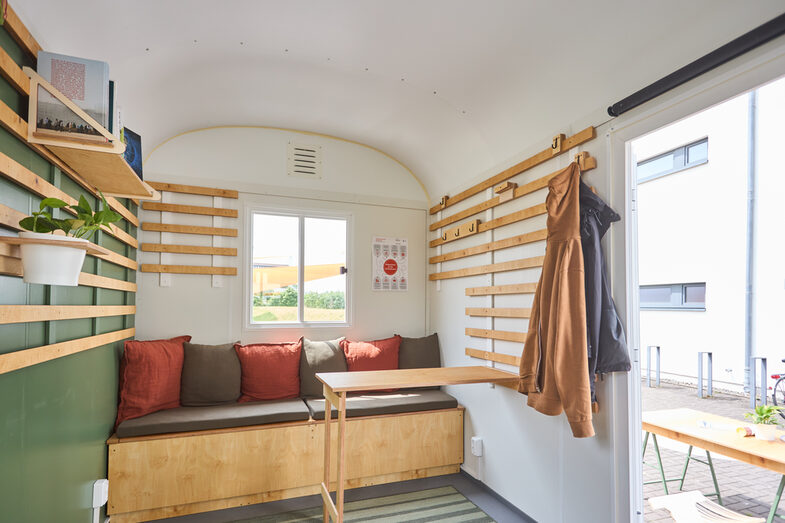 Interior shot of a converted construction trailer that serves as a mobile sustainability office. You can see a seating area with several cushions and a fold-out table. Wooden strips have been attached to the wall and are used as a coat rack.