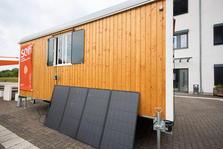 Exterior shot of a converted construction trailer with a solar panel leaning against the back. A poster on the left of the trailer reads "Soffi mobile sustainability office"