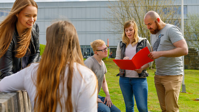 Foto von Studierenden auf dem Campus, die gemeinsam in eine Mappe schauen und sich austauschen. Im Vordergrund zwei weitere Studentinnen.__One female and tweo male students sit and stand on the campus, one student holds a folder in his hand and they exchange ideas. In the foreground two other students.
