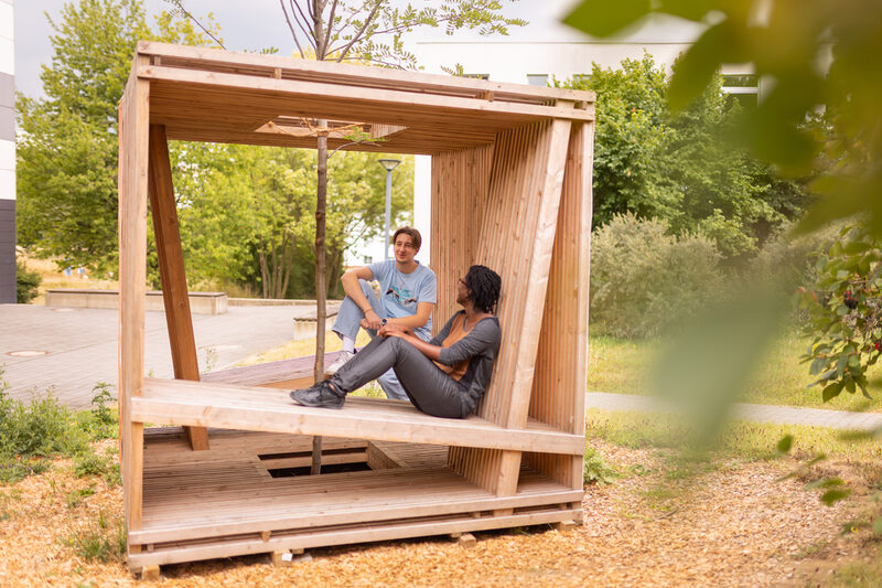 Foto von 2 Studierenden, die in einem Holzwürfel, der mit Sitzgelegenheiten ausgestattet ist, sitzen und sich unterhalten. In der Mitte des Würfels, ist ein Baum gepflanzt.