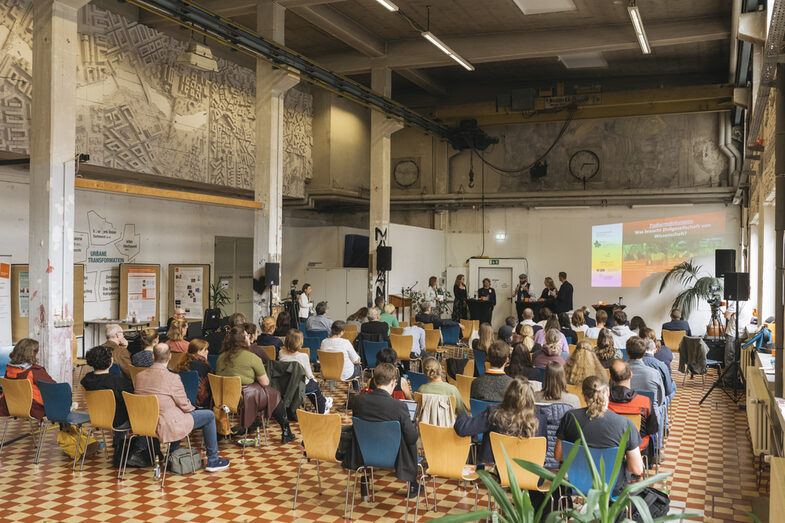 Man sieht auf dem Bild die Totale des Veranstaltungsorts Werkhalle. Es sind viele Menschen in Stuhlreihen von hinten fotografiert die einem Vortrag zu hören. Der Raum ist hoch, groß, hell