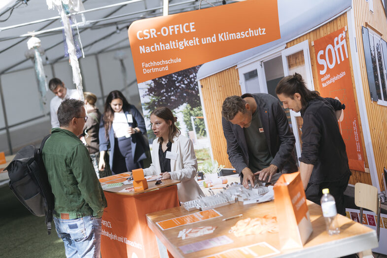 Auf dem Bild ist eine Rezeption aufgebaut. Man sieht eine Rezeptionistin im freundlichen Gespräch mit einer weiteren Person die mit dem Rücken zur Kamera steht. Im Hintergrund ist eine Werbewand aufgebaut. Dort steht "Fachhochschule Dortmund"
