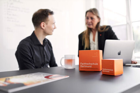 Foto von zwei Personen zusammen an einem Tisch und gucken sich gegenseitig an. Eine der Person sitzt am Tisch und die andere steht am Tisch. Auf dem Tisch steht ein Notebook sowie zwei orangene FH Würfel und eine FH Tasse.