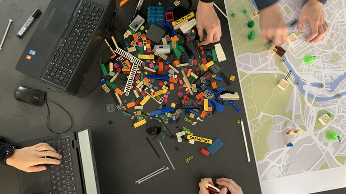 Blick von oben auf einen Tisch mit Laptops, einer Landkarte und Legosteinen in der Mitte sowie Händen von Workshop-Teilnehmenden.