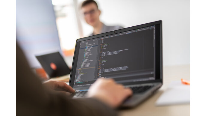 Foto eines Laptops im Closeup mit Händen an der tastatur, dahinter unscharf erkennend eine weitere Person sitzend.