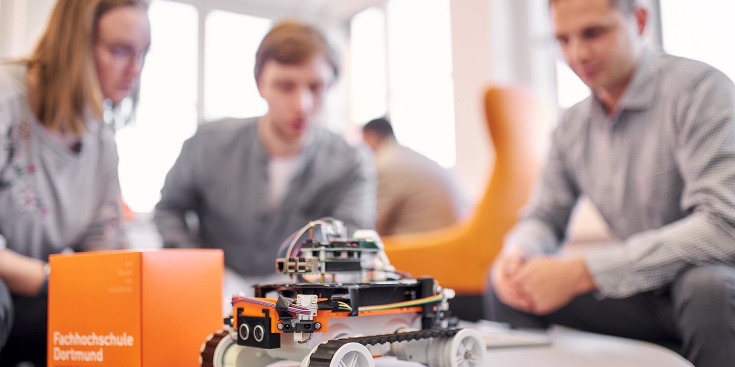 Photo of a 4-wheeled robot standing on a round table together with an orange FH cube. In the background, three people are sitting around it out of focus. They are talking about something.