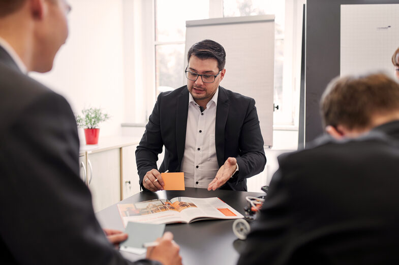 Auf diesem Foto haben sich vier Personen an einem Tisch versammelt. Bei zwei der Personen kann man erkennen, dass sie Karteikarten in der Hand halten sowie einen Kugelschreiber. Eine Person mit den Karteikarten zeigt zudem auf eine Zeitschrift und möchte dazu etwas erklären.