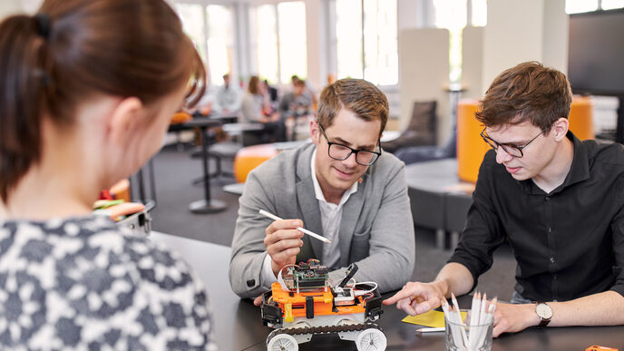 Two people are sitting at a table on which the 4-wheeled robot is standing. Both are leaning slightly forward and both are pointing towards the robot. One is pointing at something with a pencil and the other with a finger. You can also see a person standing on the opposite side of the table, attentively watching the other two people.