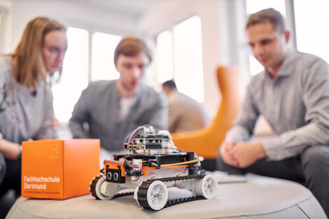 Foto von einem 4-rädrigen Roboter zusammen mit einem orangenen FH Würfel auf einem runden Tisch stehen. Im Hintergrund sitzen unscharf drei Personen drum herum. Die sich über etwas austauschen.