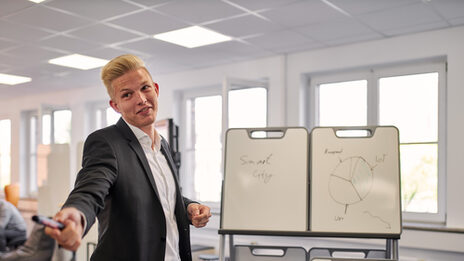 Here, a person is standing in front of a holder with three smaller whiteboards and has his arm outstretched. He has a board marker in his hand, which he apparently wants to hand over to another person so that they can write something on the whiteboards.