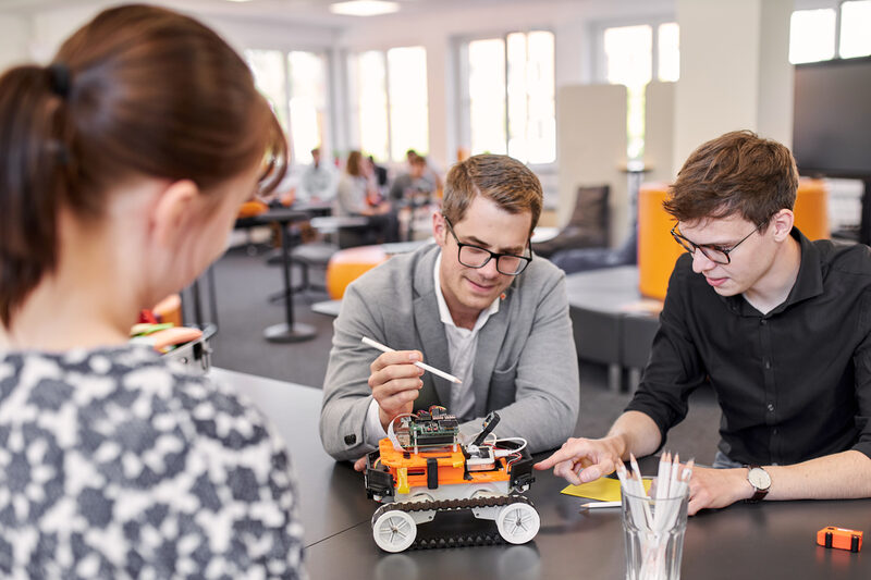 Zwei Personen sitzen an einem Tisch, worauf der 4-rädrige Roboter steht. Beide sind leicht vorgebeugt und beide zeigen zum Roboter hin. Einer zeigt mit einem Bleistift auf etwas und der andere mit dem Finger. Des Weiteren sieht man eine Person, die auf der gegenüberliegenden Seite des Tisches steht und aufmerksam den anderen beiden Personen zusieht.