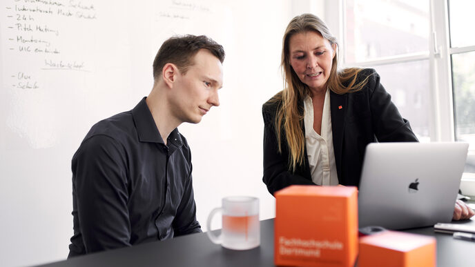 Photo of two people sitting together at a table and looking together at the screen of a notebook. One person is sitting at the table and the other is standing at the table. The person standing at the table tells the other person something. There are two orange FH cubes and an FH cup on the table.