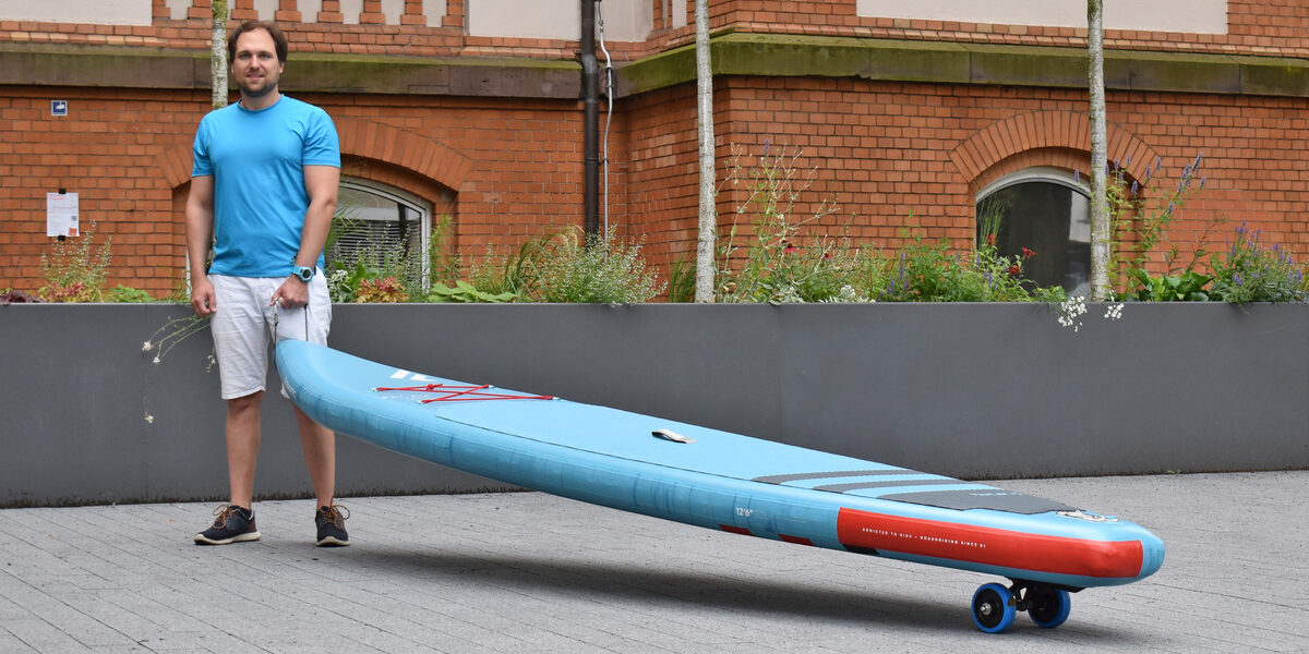 A person pulls a stand-up paddle board in front of a building with wheels attached underneath.