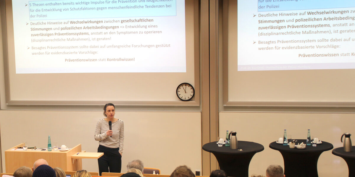 One person stands with their back to a high wall onto which a projector is projecting presentation slides. The person holds a microphone in their hand and speaks to the audience in front of them.