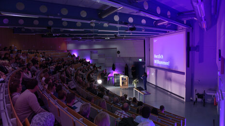 A packed lecture hall in which a woman is speaking at the front. The projection shows "Welcome!" in purple lighting.