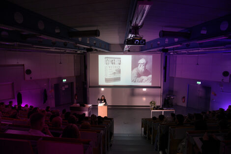 Dekanin Prof. Diana Reichle hält ihren Vortrag in einem abgedunkelten Hörsaal, während auf der Leinwand ein Buchcover und ein Porträt gezeigt werden. Der Raum ist dezent violett beleuchtet.