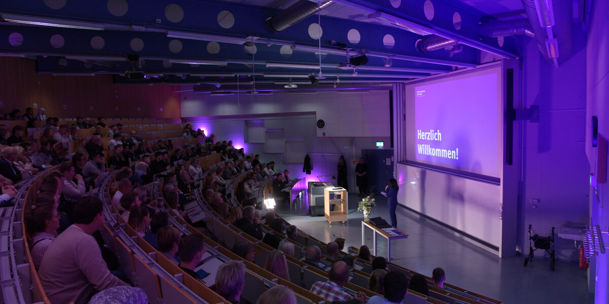 Der gefüllter Hörsaal, in dem eine Frau vorne spricht. Die Projektion zeigt „Herzlich Willkommen!“ in lila Beleuchtung.