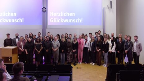Gruppenfoto der Absolvent*innen mit auf der Bühne des Hörsaals.