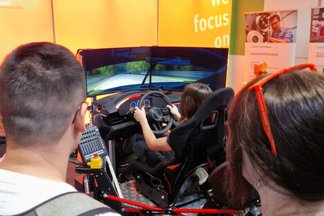 Two people watch as another person controls the driving simulator.